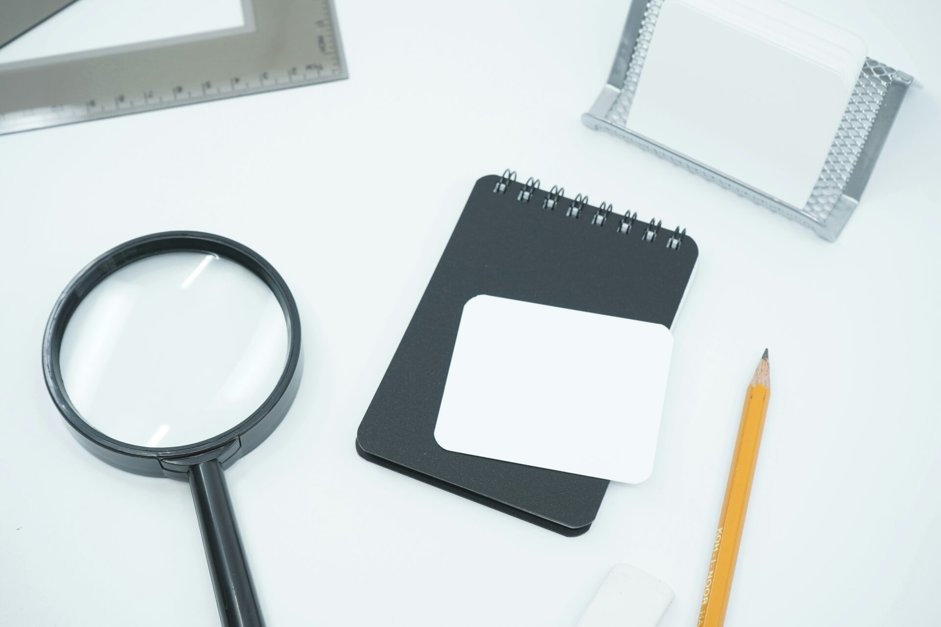 A forensic research workspace featuring a magnifying glass, pencil, and black notepad on a white desk with drafting tools, symbolizing the structural auditing and methodological hardening of academic manuscripts.